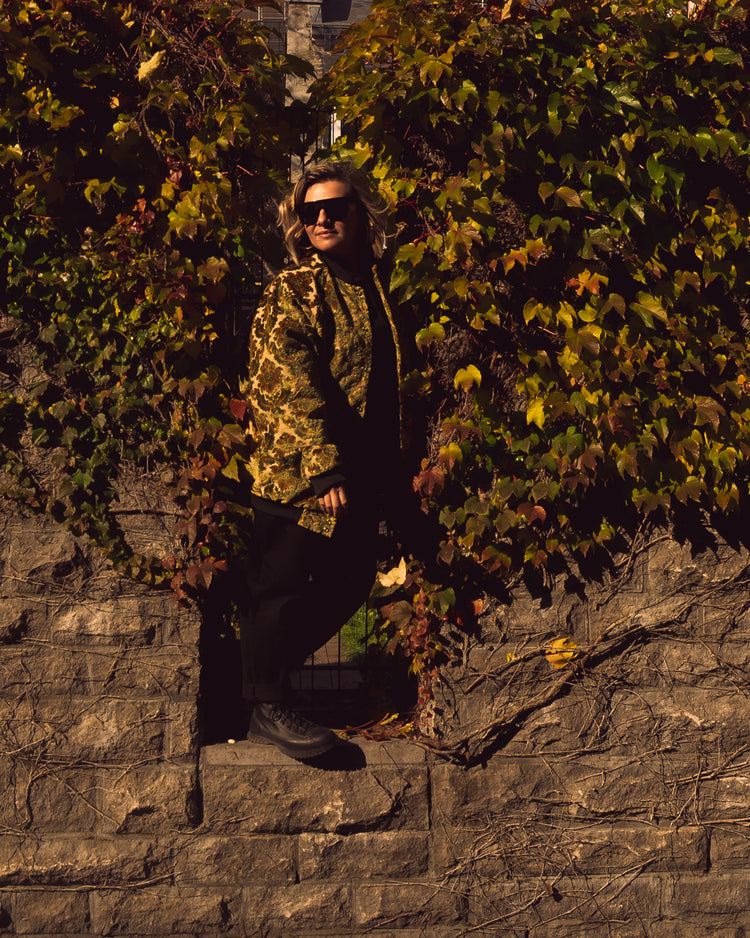 Photo of a female model wearing the House of Glass Carpetbag Bomber Jacket leaning out of a gap in an ivy covered stone wall