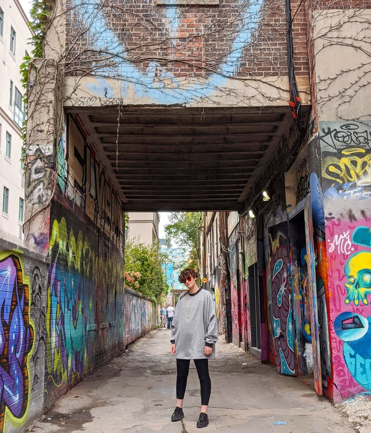picture of non binary model looking into the camera wearing house of glass cozy comfy sweater framed by graffiti covered buildings