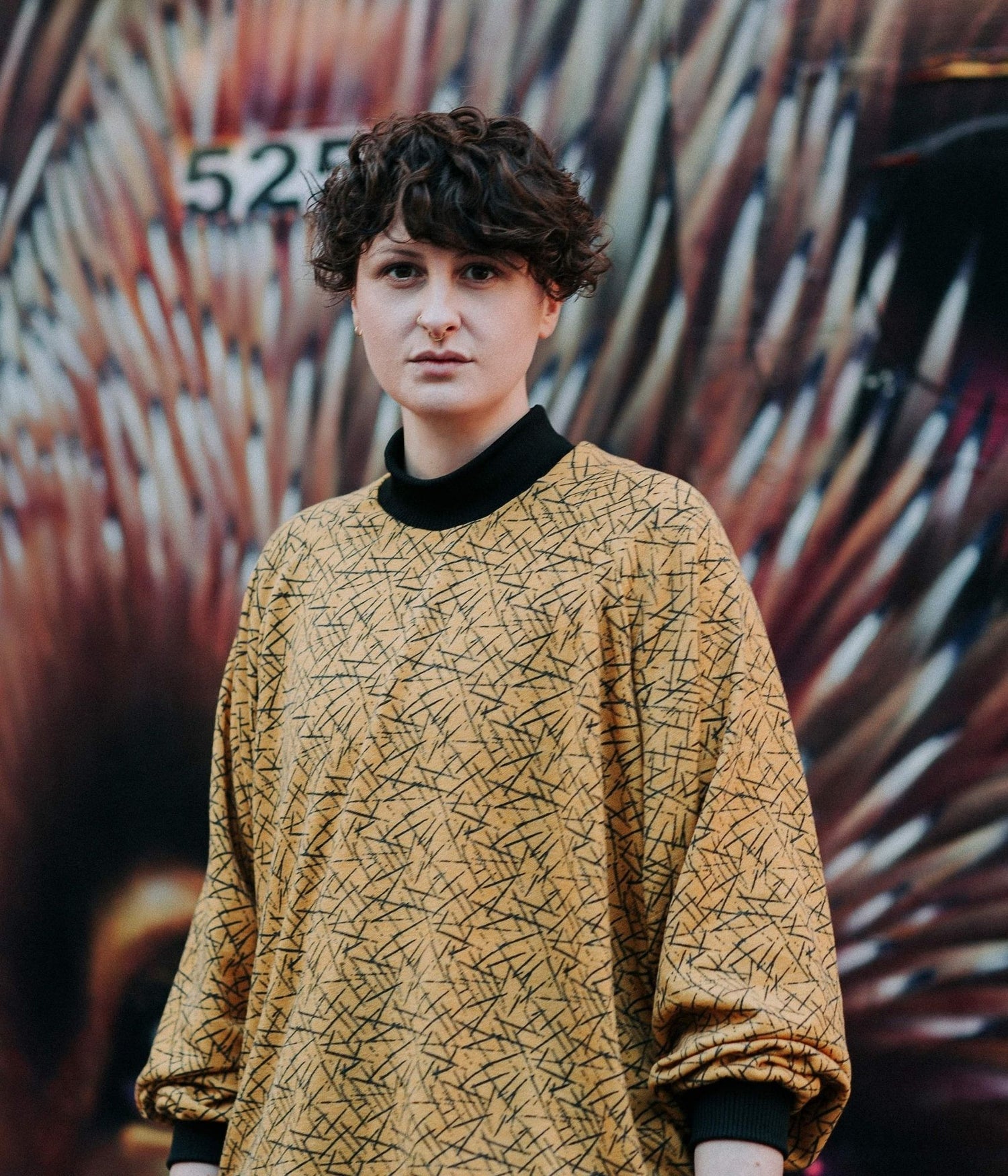 picture of non binary model looking straight at the camera wearing pickup stix jumper in front of graffiti background