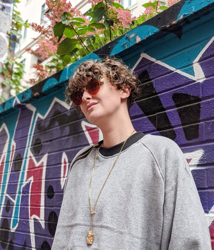 picture of non binary model wearing house of glass cozy comfy sweater standing in front of graffiti covered wall